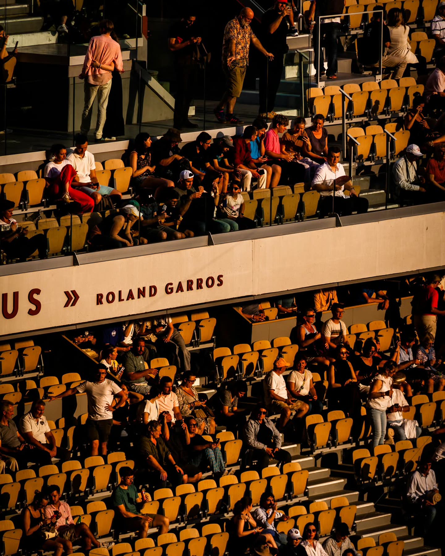 Les tribunes de Roland-Garros en fin de journée, la lumière rasante sur les sièges